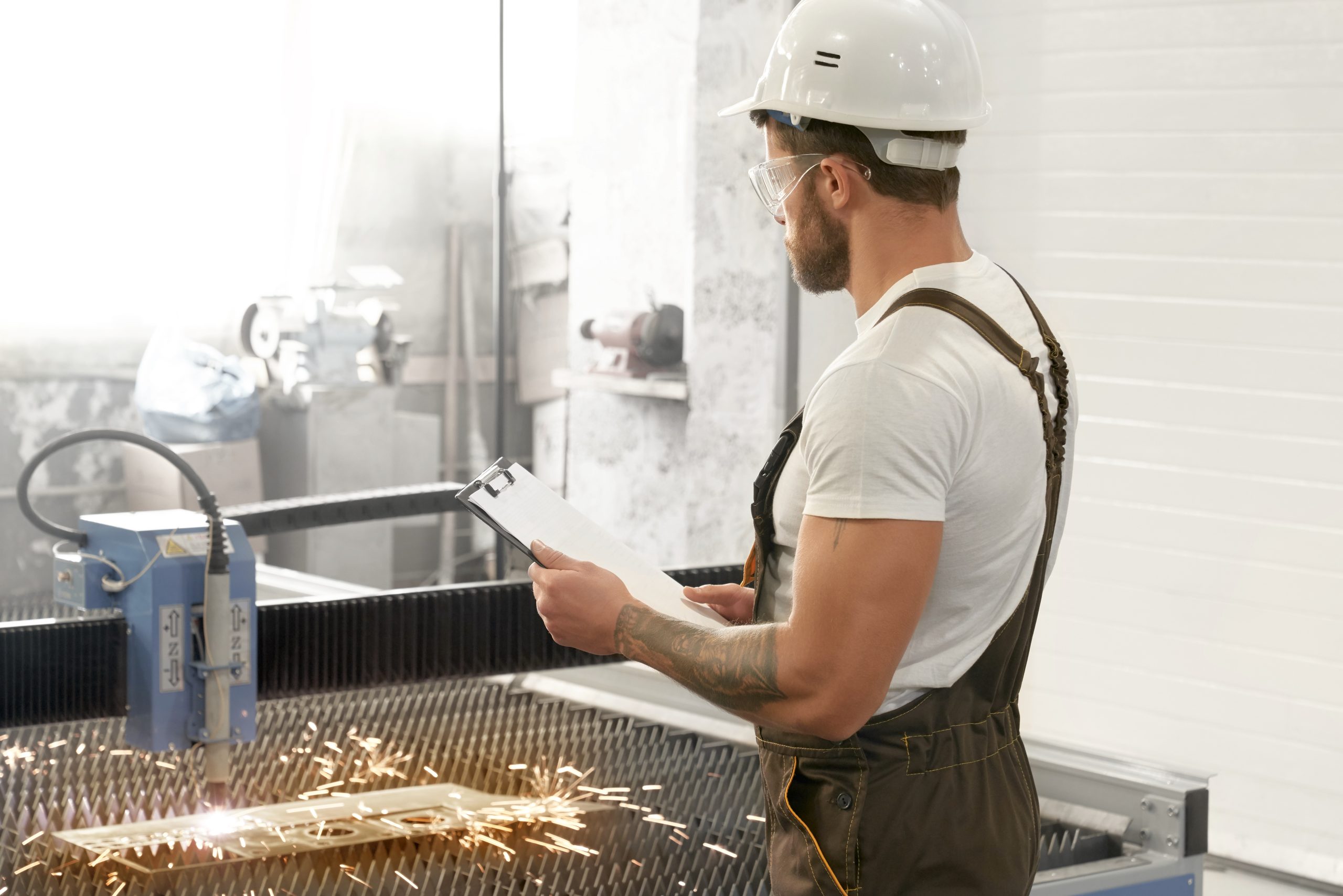 View from side of male engineer wearing uniform, protective glasses and helmet keeping folder and looking at plasma cutting. Muscular man checking process of devices and equipment.